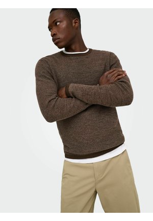 Man met gekruiste armen die een bruine gebreide trui draagt over een wit overhemd en beige broek, kijkend naar links tegen een effen achtergrond.