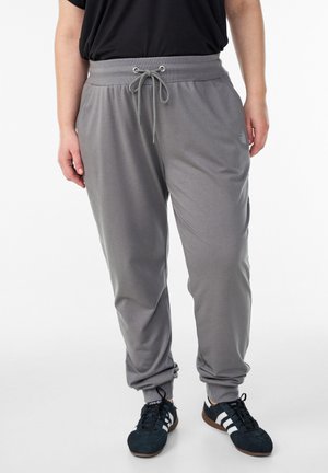 Osoba ubrana w szare spodnie dresowe ze sznurkiem oraz czarne buty sportowe z białymi paskami na białym tle.