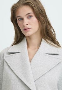 Jeune femme aux cheveux châtain clair portant un manteau en laine gris à larges revers, regardant légèrement sur le côté devant un fond clair et uni.
