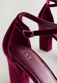 Bordeaux fluwelen hoge hakken sandalen met een vierkante hak, enkelband en logo-detail op de binnenzool. Zachte textuur en slank ontwerp.