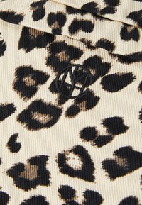 Leopardų rašto audinys su smėlio spalvos fone, turintis juodų ir rudos dėmių. Įtrauktas juodas siuvinėtis logotipas. Tekstūruotas kordūrojo medžiaga.