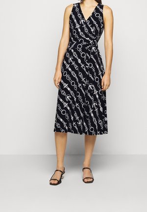 Kobieta nosząca granatową sukienkę na ramiączkach z białymi wzorami łańcuchów i kotwic, w połączeniu z czarnymi sandałami na obcasie z odkrytymi palcami.