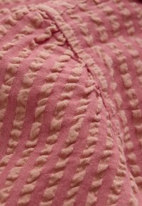 Miękki różowy materiał z podniesionymi teksturowanymi paskami i widocznym szwem, przedstawiający zbliżenie tkaniny o splocie i tuftingiem.