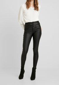Zwarte hoge taille nepleer legging met een gladde textuur, met vijf knopen aan de voorkant en twee zijzakken. Draagt een witte blouse en zwarte hakschoenen.