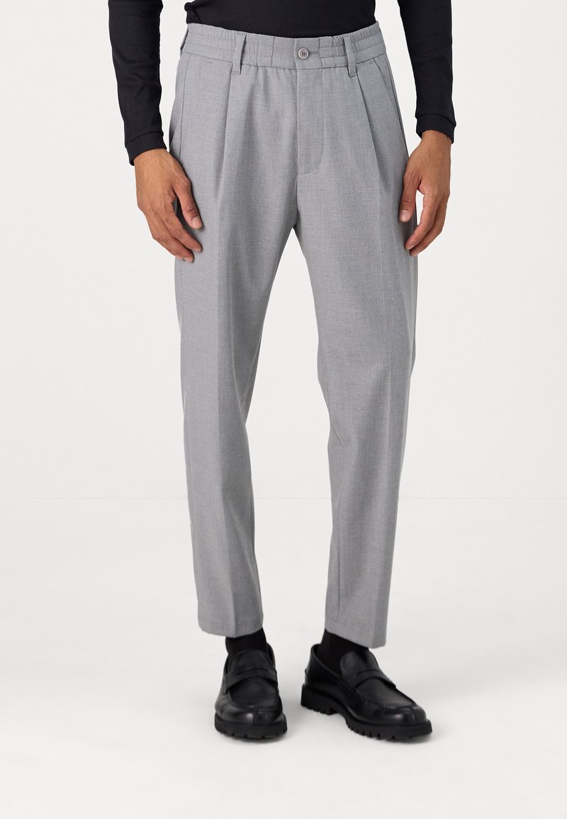 Pantalons plissés gris clair en tissu texturé, dotés d'une fermeture à bouton et d'une coupe fuselée, associés à des chaussures et des chaussettes noires.