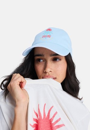 Jonge vrouw met donker haar en sproeten draagt een lichtblauwe pet met een palmboomontwerp, terwijl ze een wit shirt met een rode zonnegrafiek dicht bij haar mond vasthoudt.