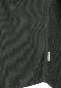 Dunkelgrüner, gerippter Cord-Stoff mit vertikalen Rillen und einem kleinen weißen Etikett mit der Aufschrift "EIGHT2NINE", das nahe einer Naht angenäht ist.