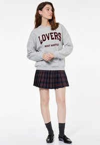 Grijze sweatshirt met "LOVERS MOST WANTED" in bordeauxrood tekst, gecombineerd met een marineblauwe en rode geruite plooienrok. Zwarte loafers en grijze sokken.