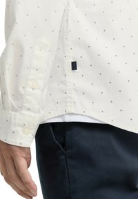 Weißes Hemd mit kleinen dunkelblauen Rautenmustern, mit Knopfmanschette und einem marineblauen Logo-Patch an der Seite. Kombiniert mit dunkelblauen Hosen.