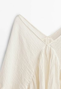 Tissu blanc texturé avec un décolleté en V et un détail froncé à l'arrière, conçu avec des drapés fluides.