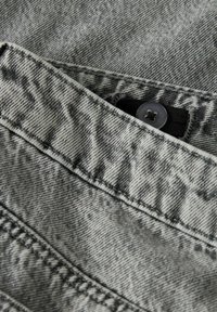 Nærbilde av grå denimstoff med synlige sømmer og en delvis skjult svart knapp festet til et svart elastisk bånd.