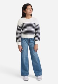 Dryžuotas megztinis baltos ir tamsiai mėlynos spalvos, tekstūra su rūbais, trumpa forma. Derinamas su šviesiai mėlynomis plačiomis kelnėmis ir baltomis sportinėmis bateliais.