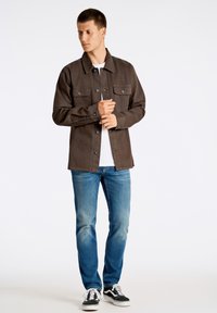 Giubbotto di jeans marrone con due tasche sul petto e chiusura a bottoni, abbinato a jeans azzurri chiari e sneakers nere. Texture liscia, design casual.