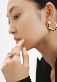Pendientes de aro en tono dorado y un gran anillo lila en una mano que toca la barbilla. El modelo tiene el cabello lacio y lleva una blusa negra.