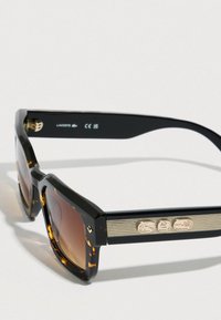 Pravokotna sončna očala z oklepnico, z rjavimi lečami z gradientom in zlatimi dekorativnimi logotipi na krakih.