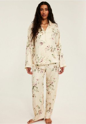 Vrouw met lang krullend haar die een crèmekleurige satijnen pyjamaset met bloemen draagt, staat blootsvoets tegen een effen lichte achtergrond.