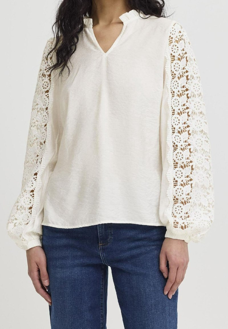 Blouse blanche avec un col en V et des manches bouffantes en dentelle ornées de motifs floraux. Texture lisse et coupe décontractée, associée à un jean bleu.