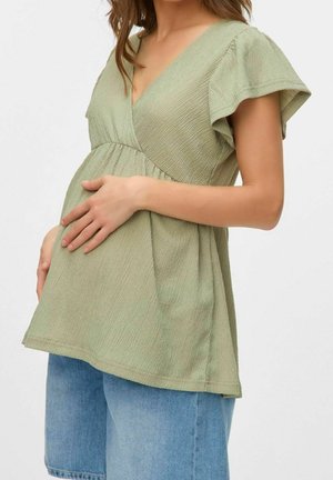 Femme enceinte portant un chemisier vert clair texturé et un short en jean bleu, tenant son ventre avec les deux mains sur un fond blanc.