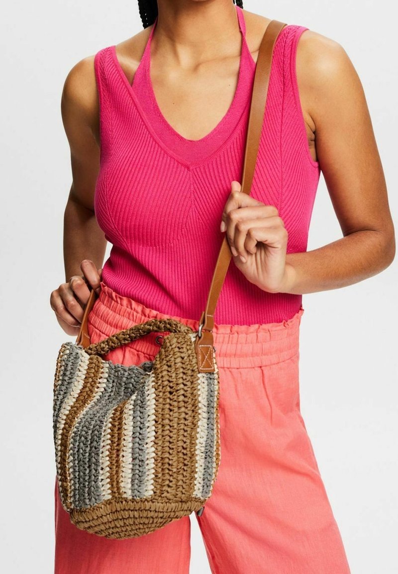Femme portant un haut sans manches rose vif et un pantalon corail, tenant un sac à main tissé rayé avec une sangle en cuir marron.