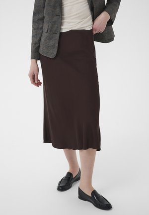 Femme portant une jupe midi marron foncé, un blazer gris, un haut blanc et des mocassins noirs, debout devant un fond uni.