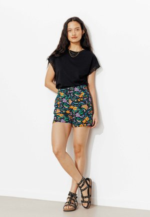 Femme debout contre un mur blanc portant un haut noir orné de dentelle, un short floral taille haute, des colliers en or superposés et des sandales noires à clous.