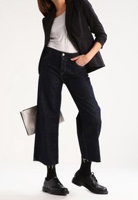 Granatowe dżinsy o szerokich nogawkach z postrzępionym wykończeniem, zestawione z szarą koszulką i czarną marynarką, uzupełnione masywnymi czarnymi butami i srebrną torebką.