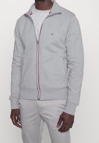 Chaqueta gris de cremallera hecha de una mezcla de algodón, cuenta con un cuello alto, acentos a rayas cerca de la cremallera y bolsillos laterales.