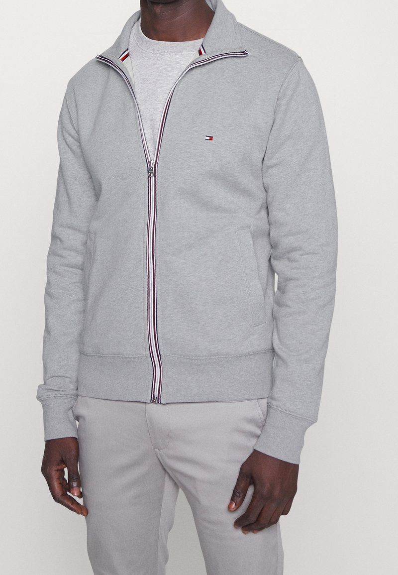 Chaqueta gris de cremallera hecha de una mezcla de algodón, cuenta con un cuello alto, acentos a rayas cerca de la cremallera y bolsillos laterales.