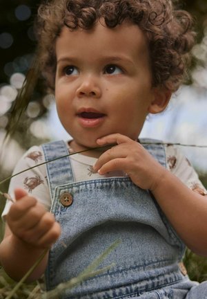 Vaikas su garbanotais plaukais, dėvintis marškinėlį su krabo raštu ir džinsinį kombinezoną, laiko žolės stiebą lauke su smalsia išraiška.