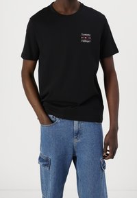 Mężczyzna w czarnej koszulce Tommy Hilfiger i niebieskich spodniach cargo, z jedną ręką w kieszeni na białym tle.