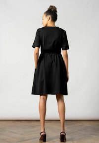 Schwarzes kurzärmeliges Kleid mit taillierter Passform und ausgestelltem Rock; verfügt über seitliche Taschen und besteht aus glattem Stoff. Rückansicht gezeigt.