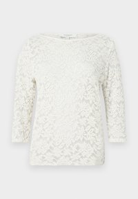 Blouse blanche à manches longues en dentelle avec motif floral, col bateau et tissu semi-transparent.