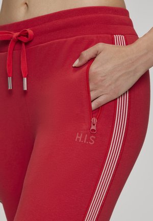 Hand in Reißverschlusstasche roter Jogginghose mit weißen Seitenstreifen und Kordelzugbund.