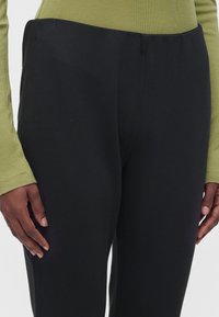 Personne portant un pantalon noir taille haute et un haut olive à manches longues, mains détendues sur les côtés, montrée de la taille jusqu'au milieu de la cuisse.
