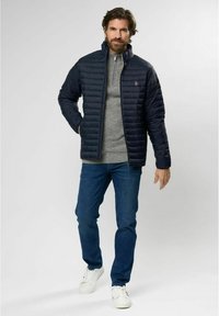 Marineblå quilted jakke med høy krage, glidelås foran og logo-detalj, paret med en grå genser, blå jeans og hvite sneakers.