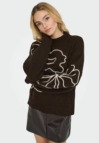Pull en tricot marron avec un motif floral blanc, doté d'un col montant et de manches surdimensionnées, associé à une mini-jupe en cuir noir.