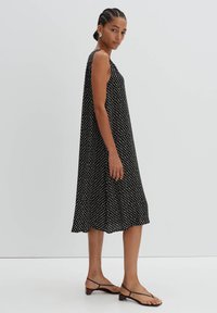 Schwarzes ärmelloses Kleid mit weißen Pünktchen, ausgestelltes Design, leichtes Material, bis zur Mitte der Waden reichend, kombiniert mit schwarzen Riemchensandalen.