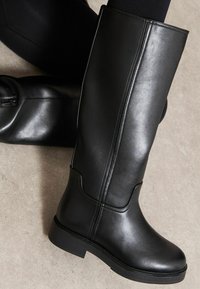 Térd fölé érő fekete műbőr csizmák, lekerekített orral, sima textúrával, minimalista dizájnnal és alacsony block sarokkal. Finom varrás részlet.