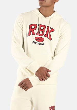 Persoon draagt een gebroken-witte Reebok-hoodie en bijpassende broek met het "RBK"-logo in rood en zwart op beide kledingstukken, handen samengevouwen voor het lichaam.