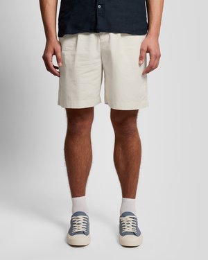 Moški, oblečen v bež kratke hlače do kolen, bele nogavice, modre superge in črno srajco s sponkami, stoji na sivem ozadju.