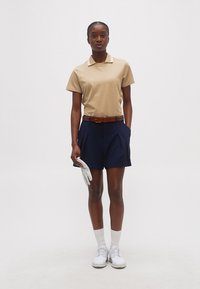 Γυναίκα που στέκεται κρατώντας ένα λευκό γάντι γκολφ, φοράει μπεζ πόλο, σκούρα μπλε σορτς, καφέ ζώνη, λευκές κάλτσες και λευκά αθλητικά παπούτσια.