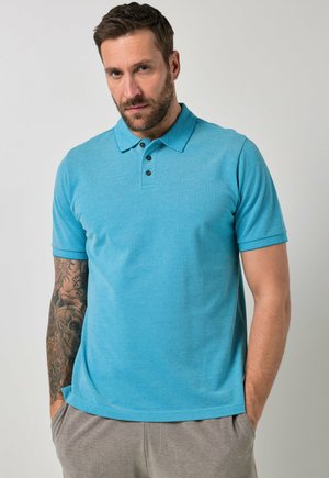 Uomo con braccio destro tatuato che indossa una polo blu a maniche corte e pantaloni grigi, in piedi con le mani nelle tasche.