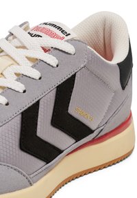 Graue Sneaker aus einer Kombination aus glattem Textil und Wildleder. Schwarze Chevron-Akzente, cremefarbene Schnürsenkel und ein rotes Außensohlen-Detail.