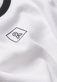 Tissu blanc avec un petit carré contour noir contenant une lettre A stylisée et un bord de col noir visible.