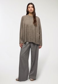 Maglione a maglia marrone chiaro con un taglio morbido e polsini a coste, abbinato a pantaloni grigi larghi. Il maglione presenta bottoni laterali come dettaglio.
