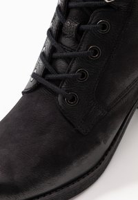 Bota de cuero negra hasta el tobillo con acabado texturizado, punta redonda, seis ojales metálicos con cordones negros y un ligero tacón. Vista en primer plano.