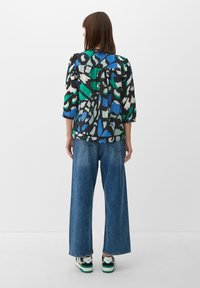 Blouse multicolore avec une base noire, présentant des motifs abstraits verts et bleus, à manches 3/4 et coupe ample, accompagnée de jeans ample.