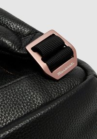 Esquina de bolso de cuero negro con una hebilla metálica de oro rosa y cinta negra. Texto "Maserati" grabado en la hebilla. Detalles de superficie texturizada.