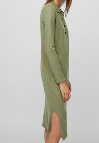 Femme portant une robe mi-longue à manches longues de couleur vert olive, avec un col, des boutons noirs et une fente latérale, sur un fond uni gris clair.