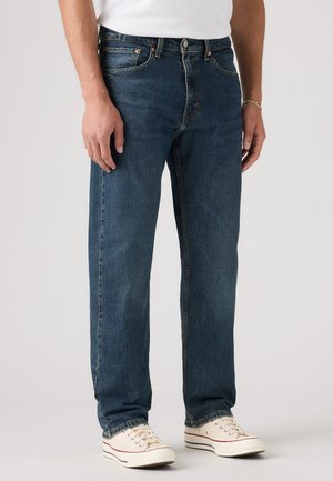 Blauwe spijkerbroek met een recht model, vijf-pocket ontwerp en koperen nieten. Gekleed met crèmekleurige sneakers.